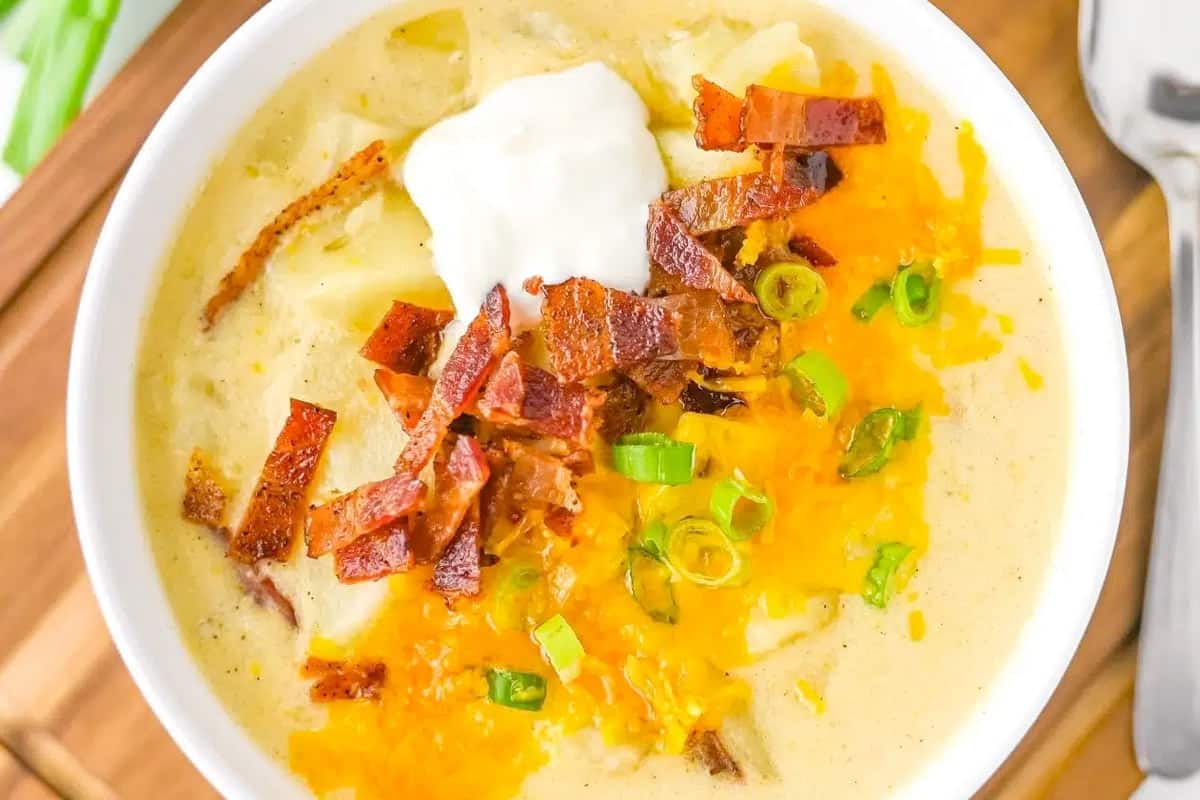 A bowl of creamy soup topped with shredded cheese, crispy bacon pieces, chopped green onions, and a dollop of sour cream, with a spoon beside the bowl.