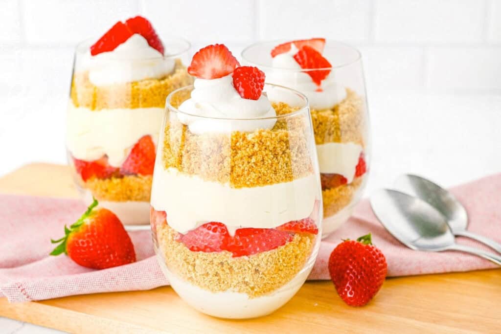 Three glass cups filled with layered cheesecake mousse, graham cracker crumbs, and fresh strawberries, topped with whipped cream and sliced strawberries, on a wooden board.