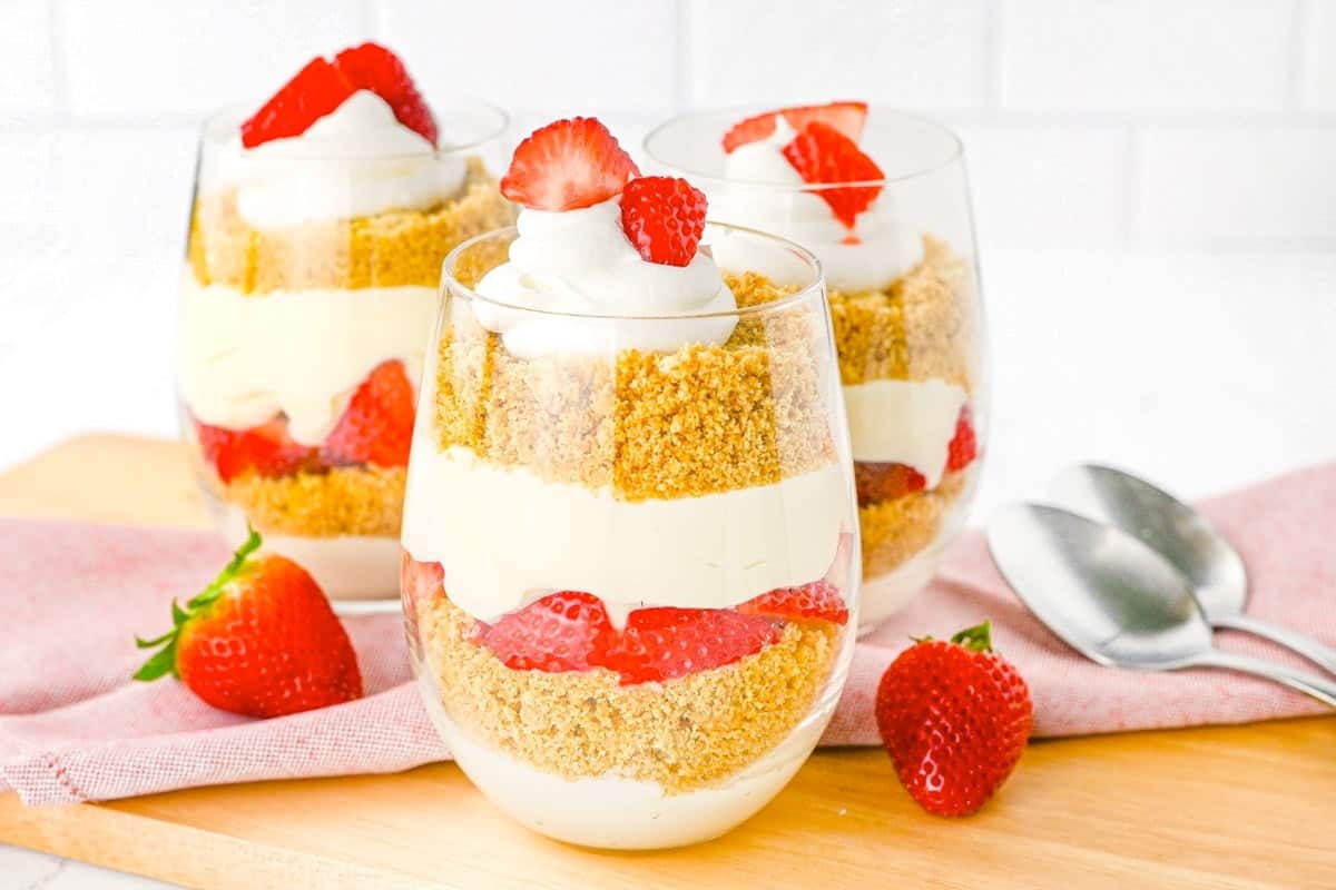 Three glass cups filled with layered cheesecake mousse, graham cracker crumbs, and fresh strawberries, topped with whipped cream and sliced strawberries, on a wooden board.