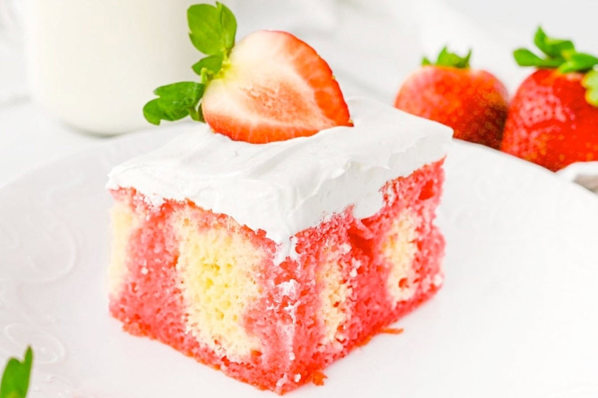 A slice of strawberry poke cake with white frosting and a halved strawberry on top, served on a white plate with fresh strawberries in the background.