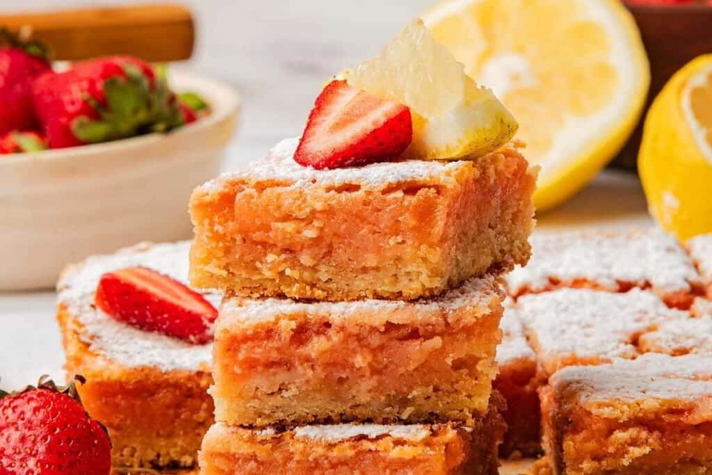 Three stacked lemon bars topped with powdered sugar, a strawberry slice, and a lemon wedge make perfect Valentines treats, with more lemon bars, lemons, and strawberries in the background.