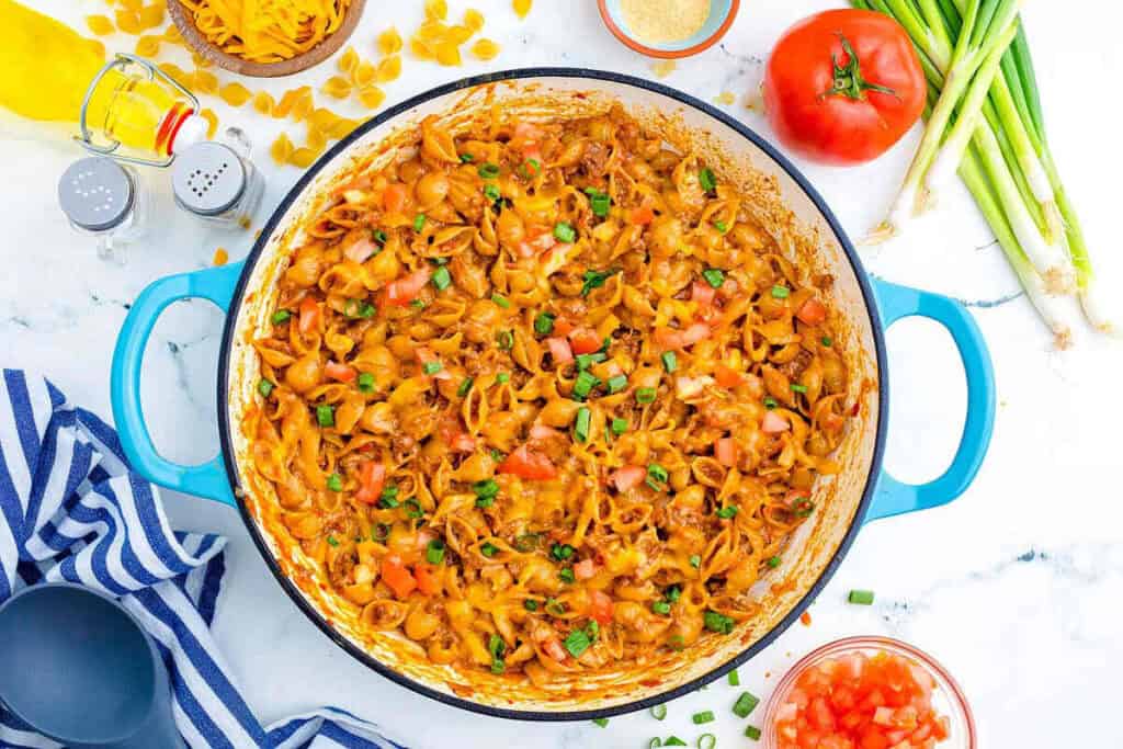 A blue pot filled with cheesy pasta topped with diced tomatoes and green onions, surrounded by fresh ingredients and kitchen utensils on a marble countertop.