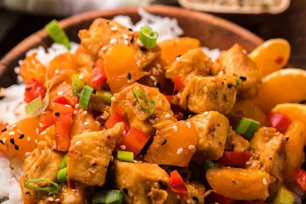 A bowl of white rice topped with chunks of chicken, orange segments, diced red bell peppers, green onions, and sesame seeds.