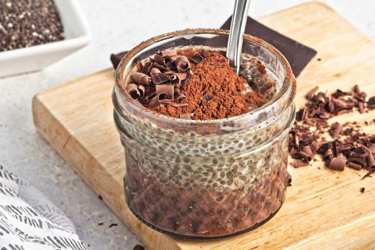 A glass jar filled with chia pudding topped with cocoa powder and chocolate shavings sits on a wooden board with a spoon inside.