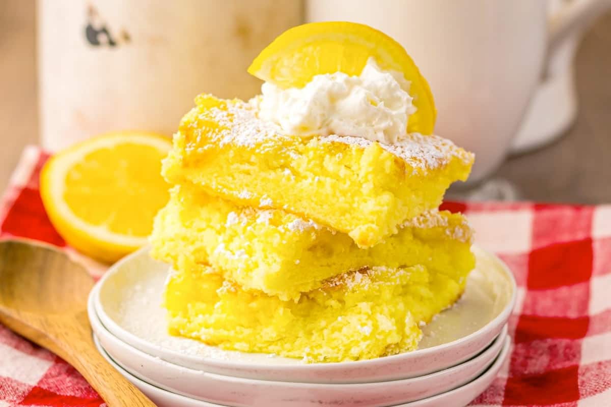 Three stacked lemon bars topped with whipped cream and a slice of lemon are served on white plates, with a wooden spoon and red checkered cloth nearby.