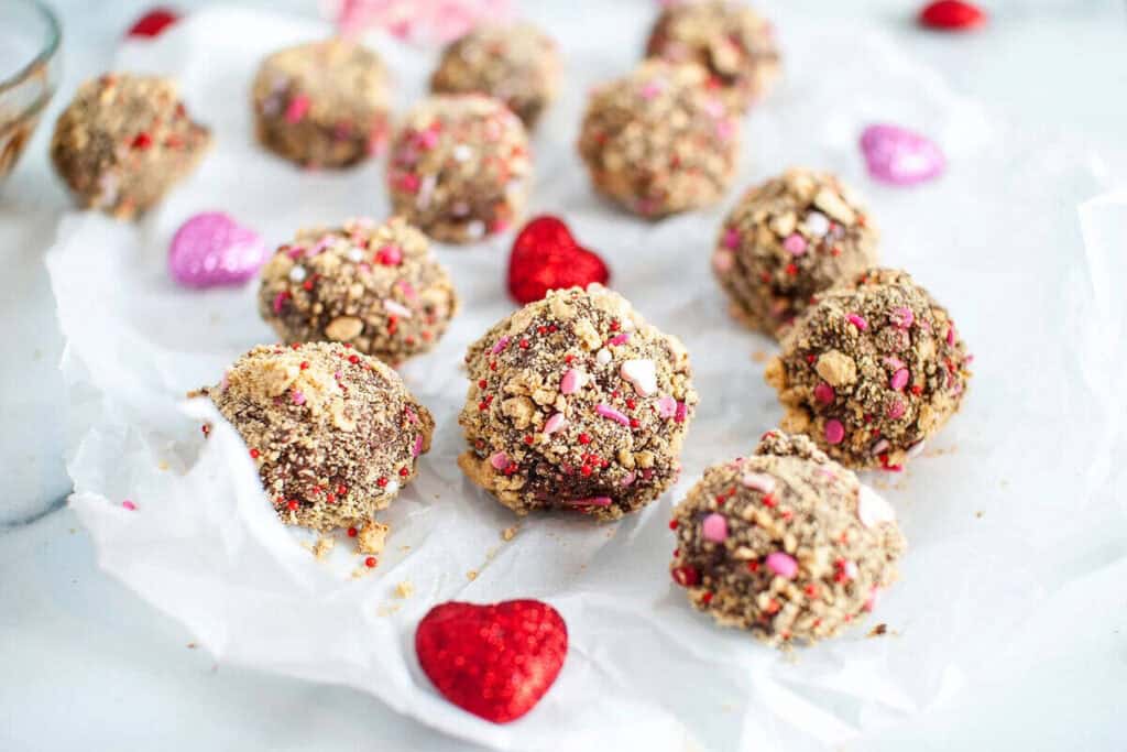 Several chocolate truffles coated in crushed cookies and colorful sprinkles are arranged on parchment paper with decorative red and pink heart shapes, making them perfect Valentine's treats.