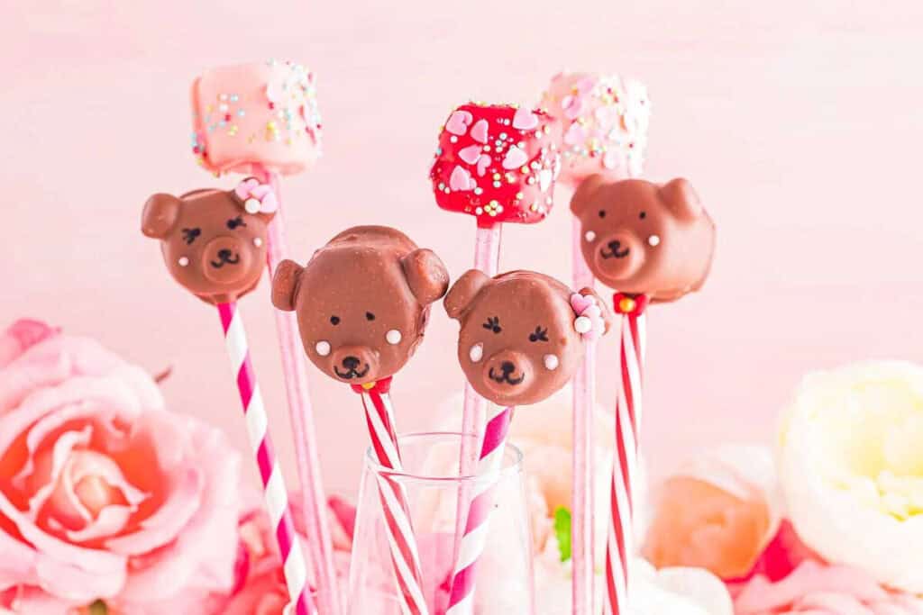 Chocolate bear and sprinkle-covered marshmallow pops on striped sticks are arranged in a glass, making adorable Valentines treats, with pink and white flowers in the background.