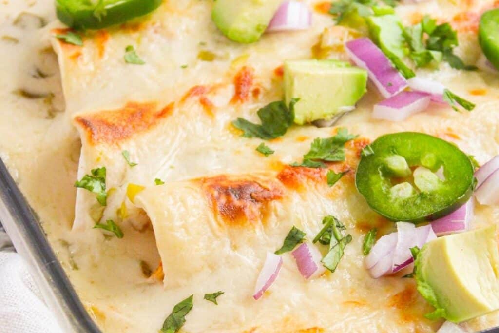 Close-up of baked enchiladas covered in melted cheese, topped with sliced jalape&ntilde;os, avocado, red onion, and chopped cilantro.