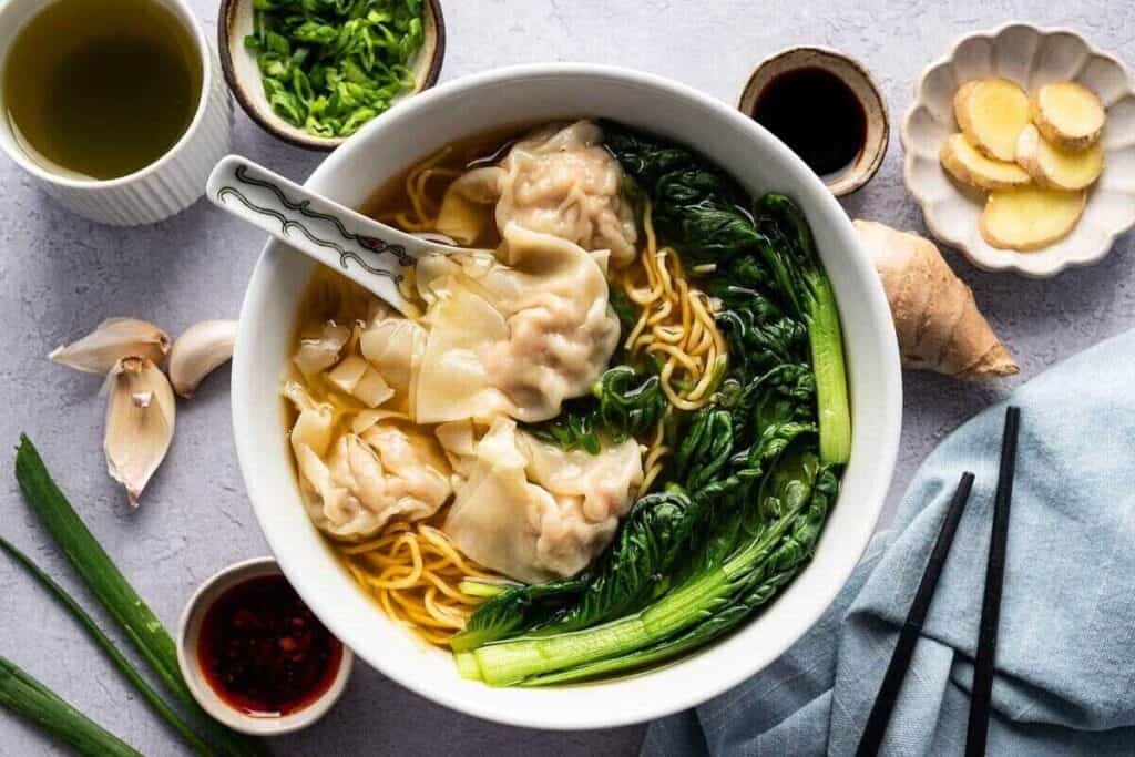 A bowl of wonton noodle soup with green vegetables, surrounded by small dishes of ginger, chili oil, chopped green onions, garlic, and tea&mdash;classic Asian-inspired dishes&mdash;with chopsticks placed nearby.