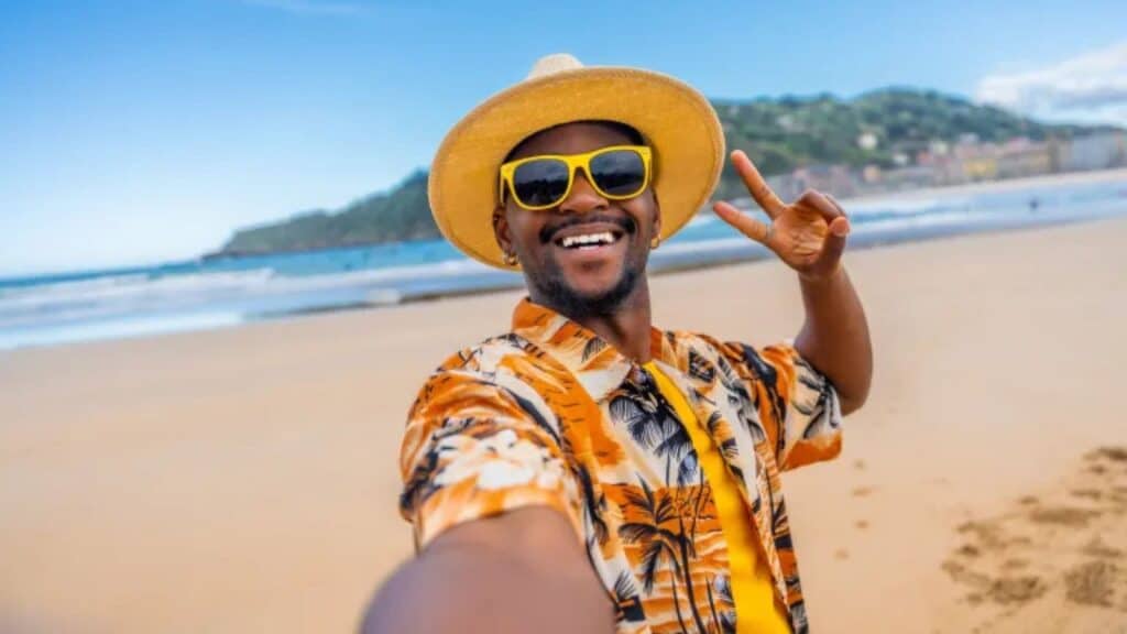 A man wearing a straw hat, yellow sunglasses, and a floral shirt takes a selfie on a sandy beach, holding up a peace sign with the ocean and hills in the background.