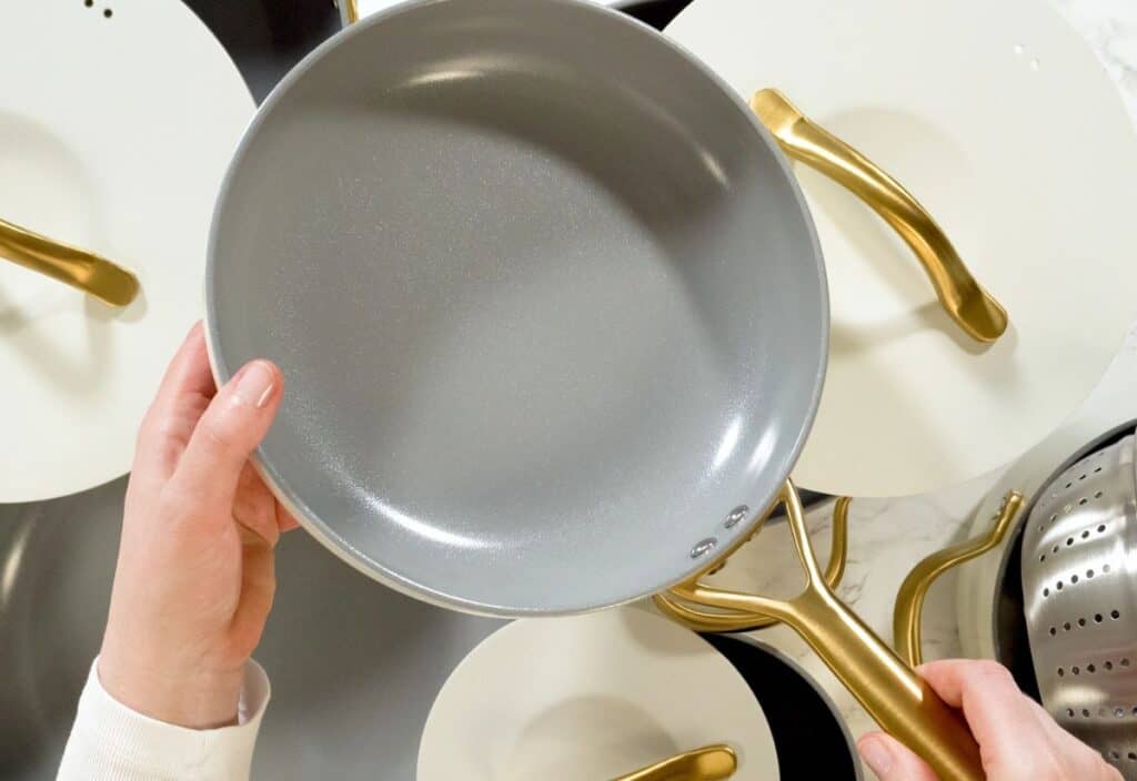 A person holds a grey nonstick frying pan with a gold handle over several similar pans arranged on a white surface.