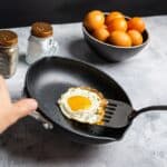 A person uses a spatula to lift a fried egg from a nonstick pan, with a bowl of eggs and salt and pepper shakers in the background.