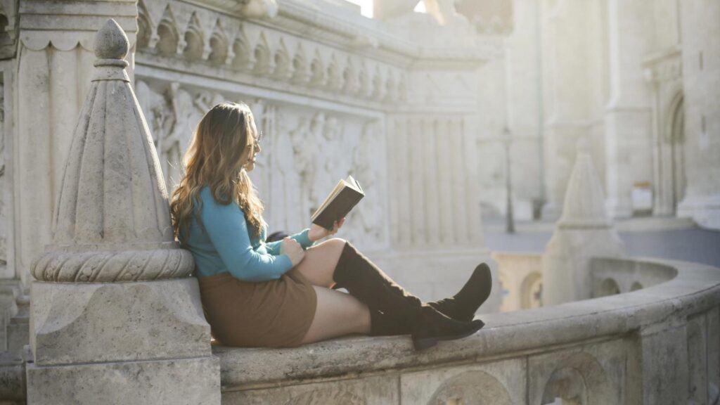 Woman with long hair, wearing a blue top, brown skirt, and black boots, sits on a stone railing outdoors, reading a book in sunlight.