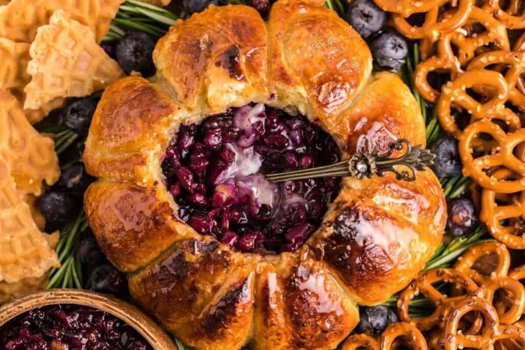 Baked pastry filled with blueberry compote, surrounded by pretzels, waffle cookies, and fresh blueberries, with a decorative spoon in the filling.