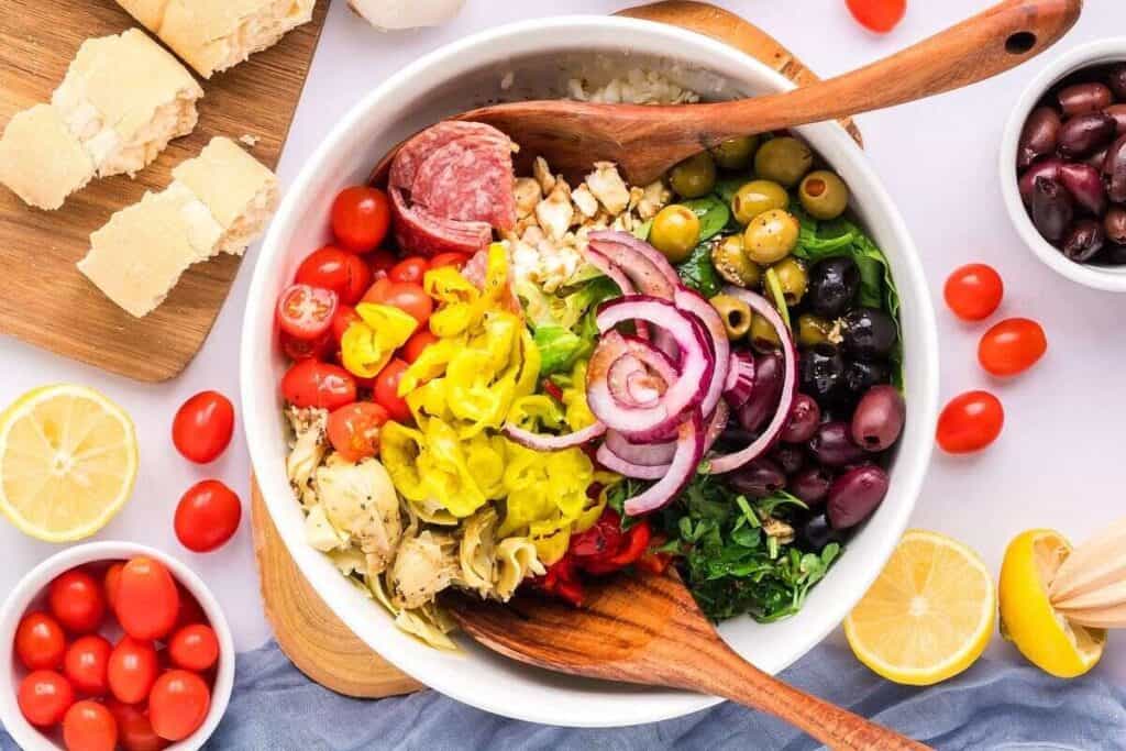 A large bowl filled with salad ingredients including cherry tomatoes, red onion, olives, salami, pepperoncini, artichoke hearts, green leaves, and cheese, with bread and lemon on the side.