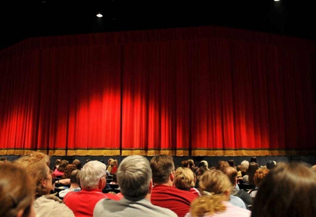 An audience of various ages and backgrounds seated in a theater, facing a large red curtain, ready for a live performance or show.