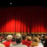 An audience of various ages and backgrounds seated in a theater, facing a large red curtain, ready for a live performance or show.