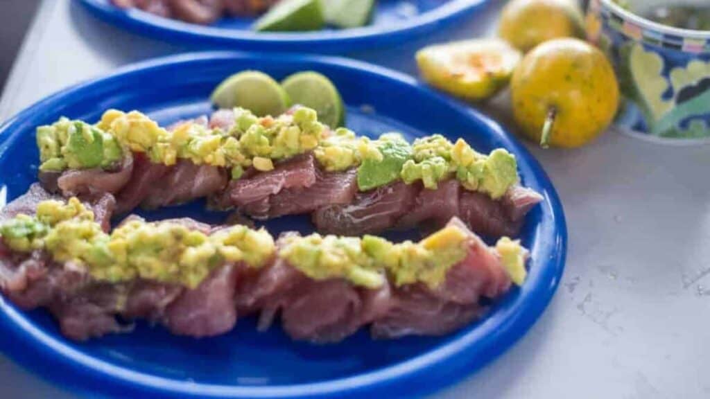 Slices of raw fish topped with diced avocado are arranged on a blue plate, with lime wedges and yellow fruit in the background.