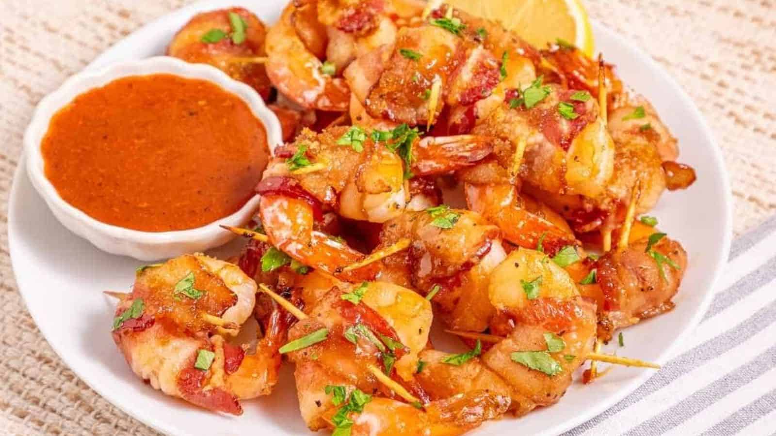 A plate of bacon-wrapped shrimp garnished with chopped herbs, served with a small bowl of reddish dipping sauce and a lemon wedge.