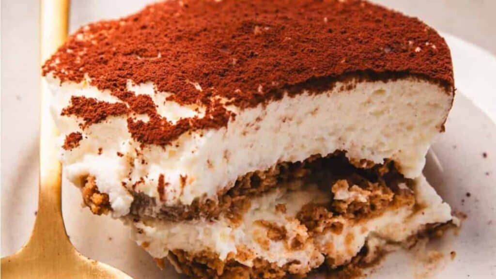 A close-up of a serving of tiramisu on a white plate, showing layers of cream, soaked ladyfingers, and a dusting of cocoa powder on top. A gold spoon is beside it.