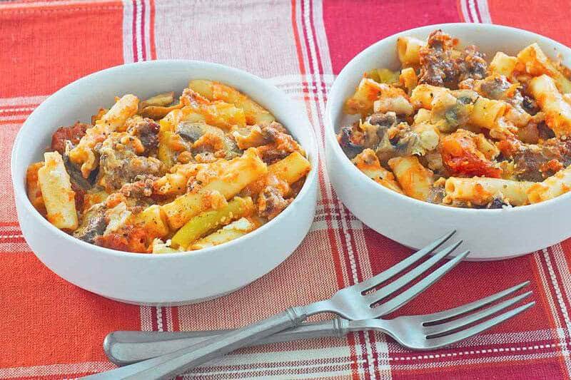 Two white bowls filled with baked pasta, ground meat, and melted cheese sit on a red plaid tablecloth next to two metal forks.