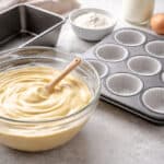 A glass bowl of cake batter with a spatula, a muffin tin lined with paper cups, a loaf pan, eggs, flour, and other baking ingredients on a kitchen counter.
