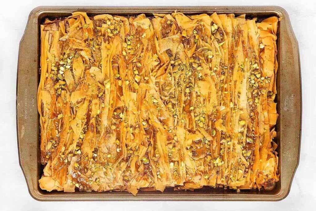 A rectangular baking tray filled with golden brown, flaky pastry topped with chopped pistachios and syrup.