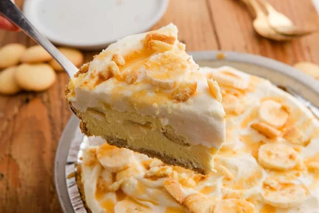A close-up of a slice of banana cream pie with whipped topping and cookie pieces being lifted from the whole pie on a wooden table.