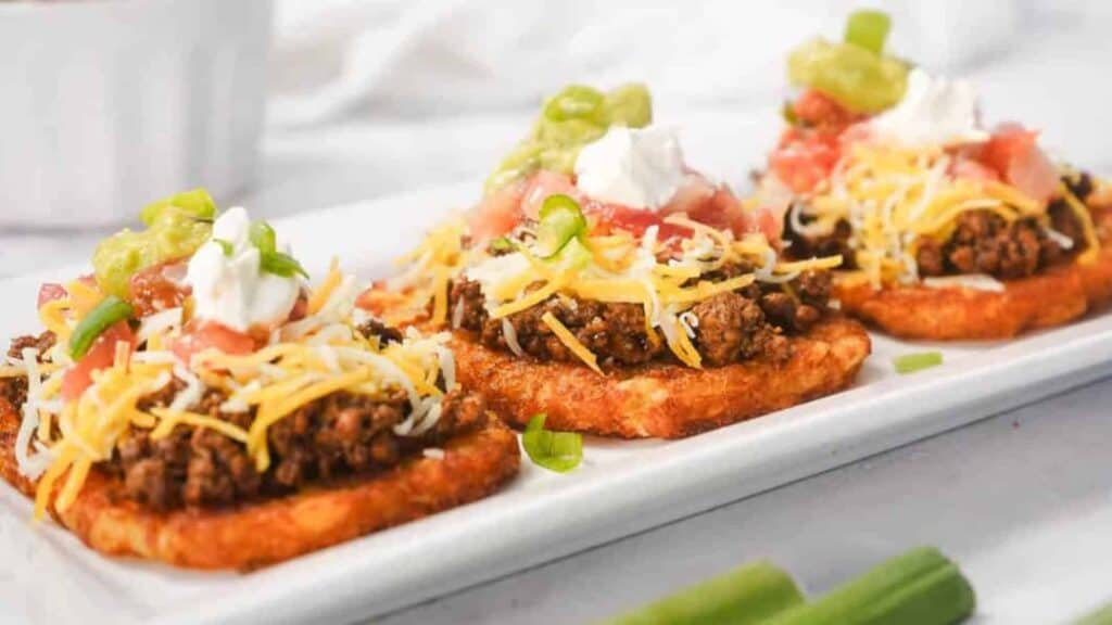 Three hash brown patties topped with seasoned ground beef, shredded cheese, diced tomatoes, sour cream, guacamole, and chopped green onions on a white rectangular plate—perfect for steady budget dinners.