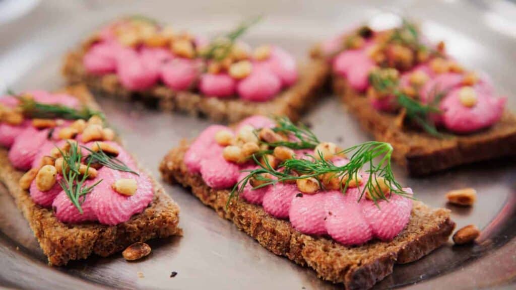 Rectangular pieces of brown bread topped with pink spread, toasted pine nuts, and sprigs of fresh dill, arranged on a metallic plate.