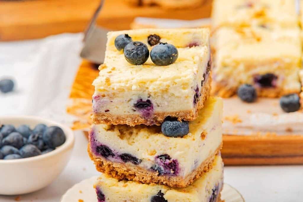Three squares of blueberry cheesecake bars stacked on a plate, topped with fresh blueberries. A bowl of blueberries and more cheesecake bars are in the background.