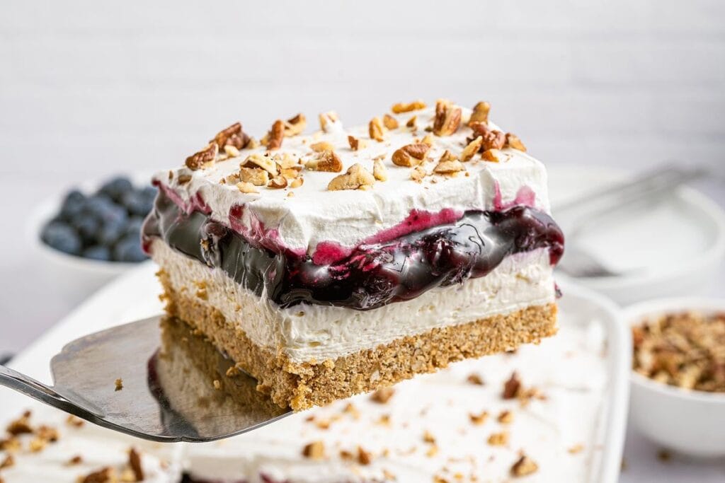 A square slice of layered dessert with a graham cracker crust, creamy filling, blueberry topping, whipped cream, and chopped pecans on a spatula.