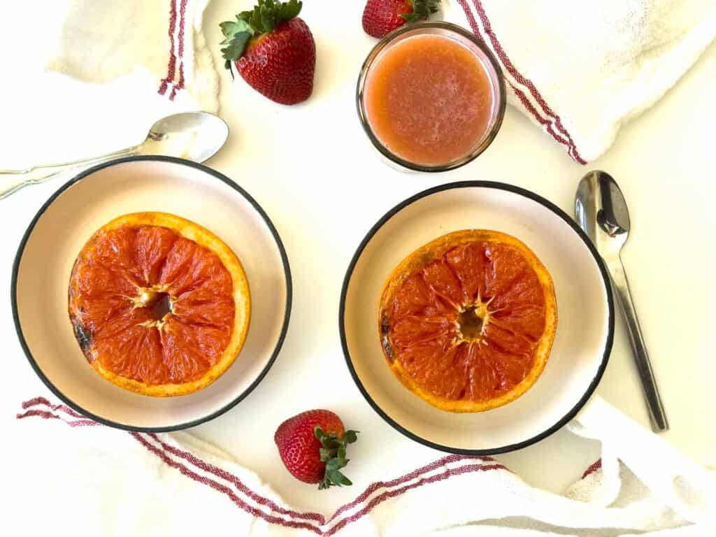 Two plates each with half a broiled grapefruit, a glass of grapefruit juice, three strawberries, two spoons, and white towels with red stripes on a white surface.