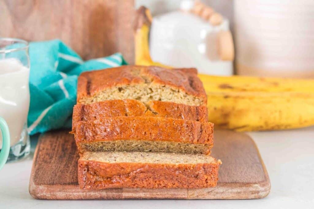 A sliced loaf of banana bread sits on a wooden board, with bananas and a glass of milk in the background.