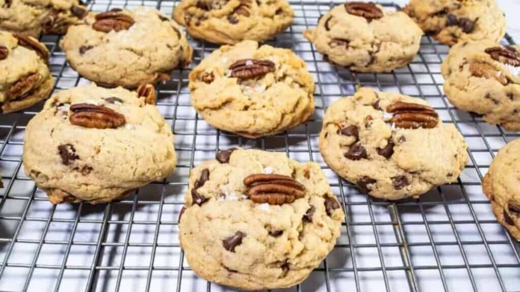 Chocolate chip cookies topped with pecan halves are cooling on a wire rack.