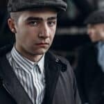 Two young men in vintage-style clothing and flat caps; one stands in focus facing the camera, while the other is blurred in the background, evoking the perfect vibe for a Peaky Blinders watch party.