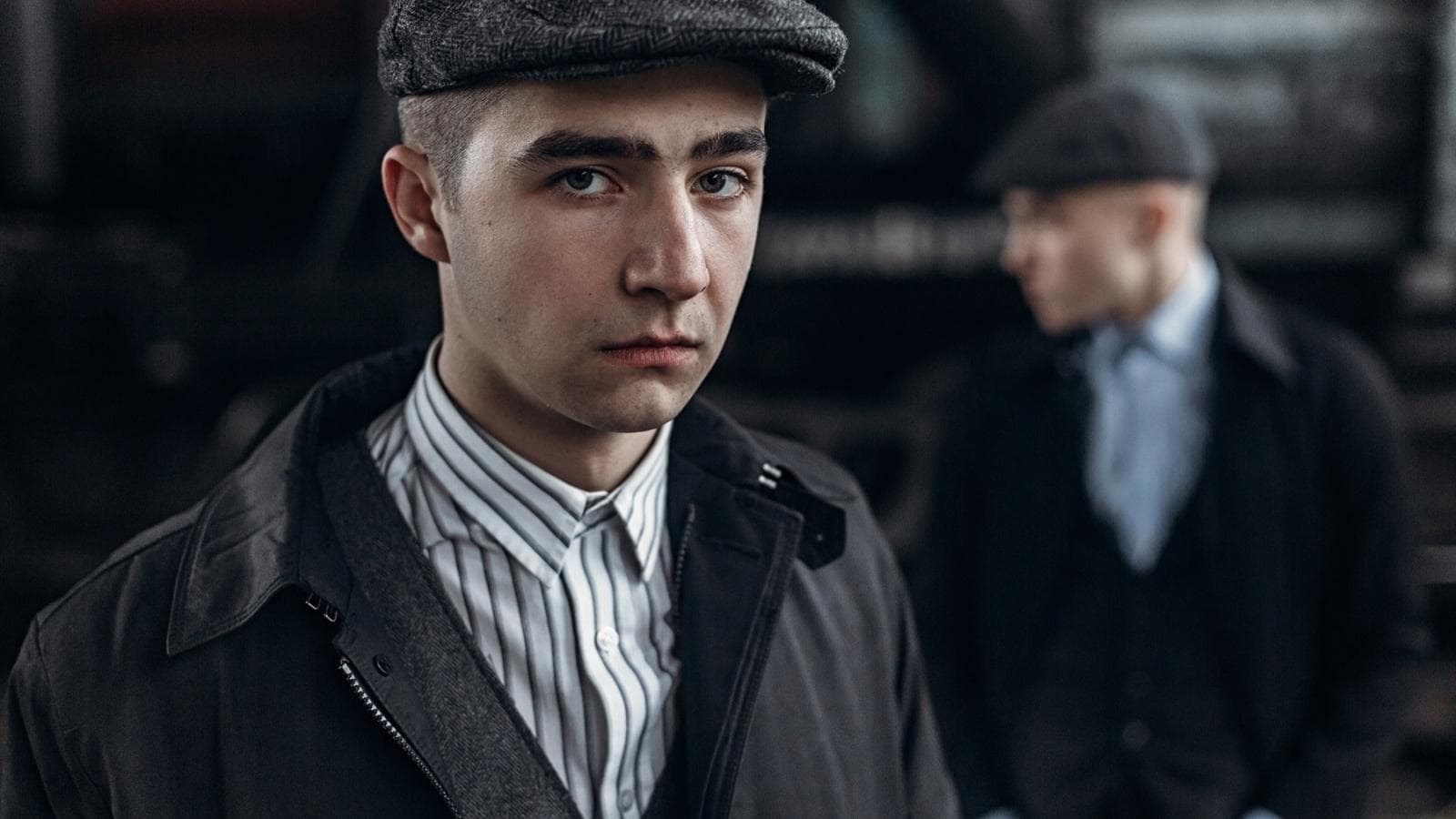 Two young men in vintage-style clothing and flat caps; one stands in focus facing the camera, while the other is blurred in the background, evoking the perfect vibe for a Peaky Blinders watch party.