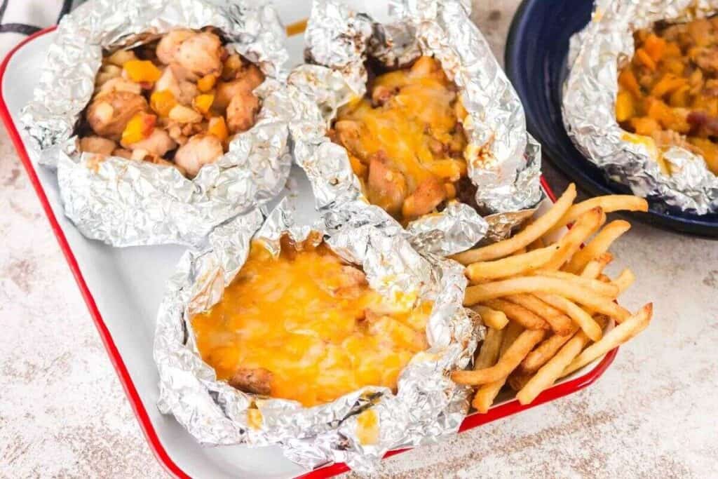 Three foil packets filled with diced chicken, vegetables, and melted cheese sit on a tray next to a serving of French fries.