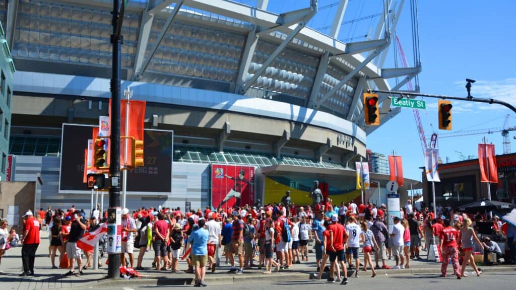 A crowd of people in red and white gather outside a large stadium on a sunny day, near an intersection with 