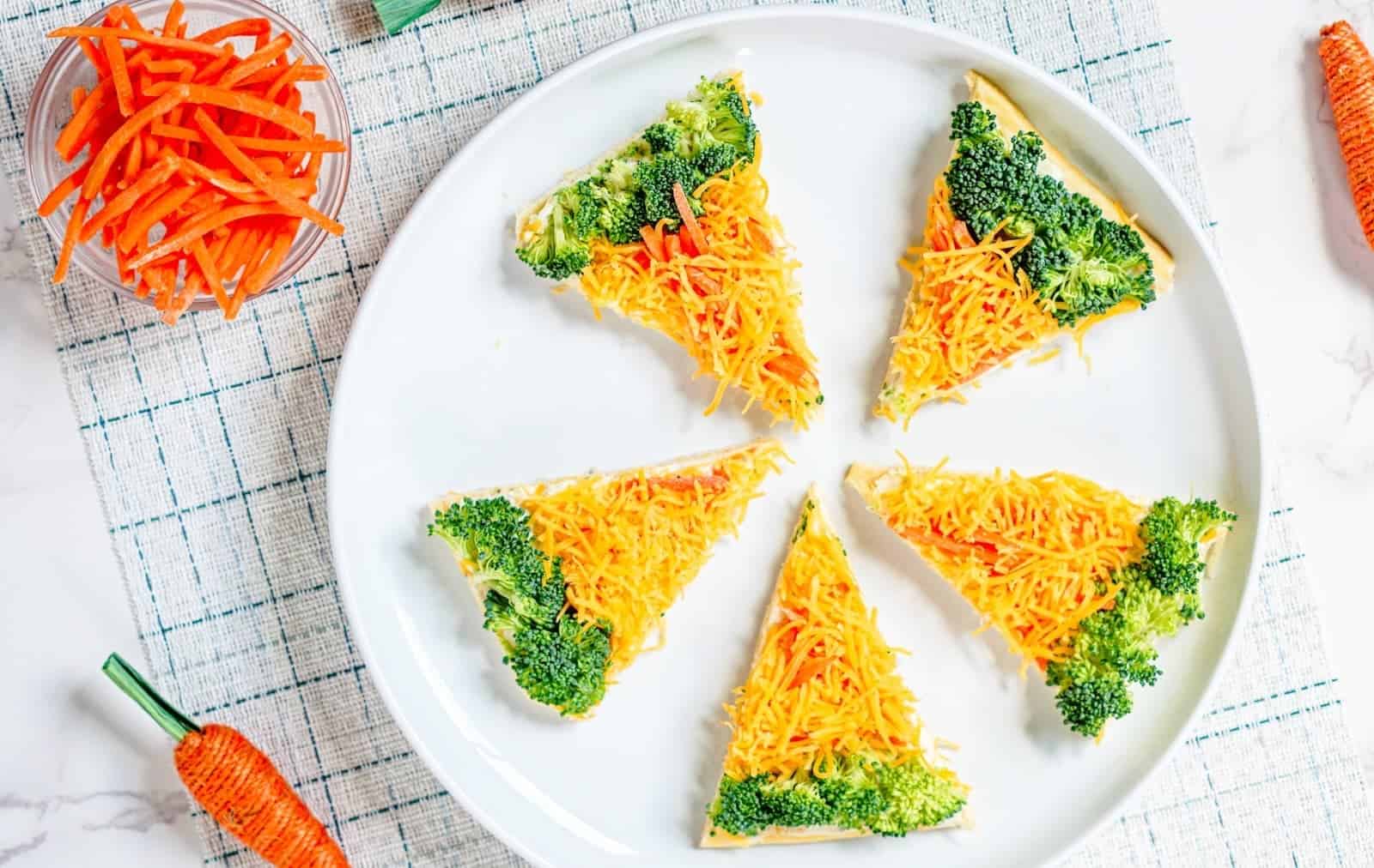 A slice of veggie pizza topped with shredded cheddar cheese, broccoli florets, and red bell pepper on a square white plate.