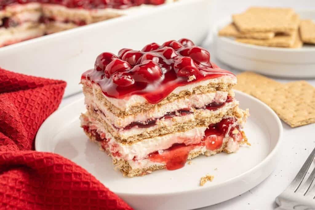 A slice of cherry layered dessert with graham crackers, cream, and cherry topping on a white plate next to a red napkin and graham crackers.