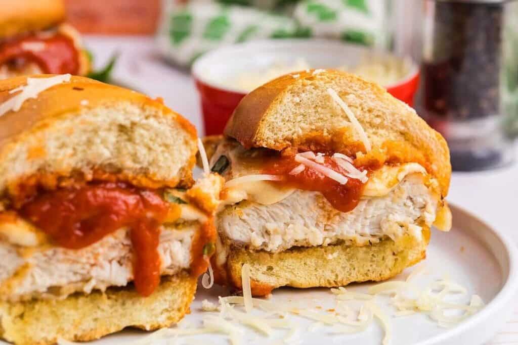 A close-up of a chicken parmesan sandwich cut in half, with melted cheese and marinara sauce on a white plate.