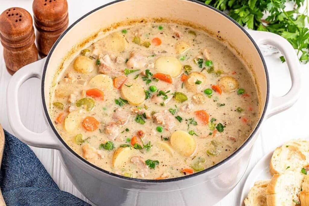 A pot of creamy chicken stew with potatoes, carrots, celery, peas, and herbs sits on a table next to sliced bread, parsley, and salt and pepper shakers.