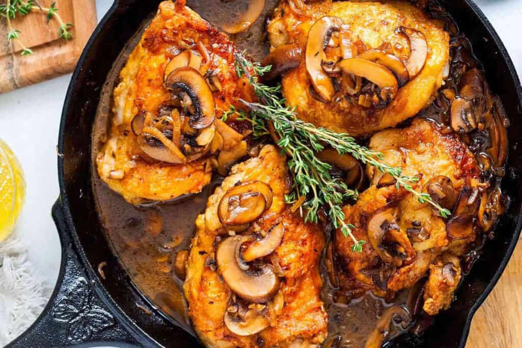 Four cooked chicken thighs topped with saut&eacute;ed mushrooms and fresh thyme in a cast iron skillet with a brown sauce.