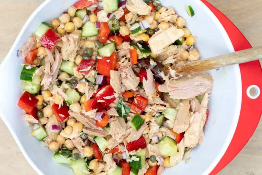A bowl of tuna salad with chickpeas, chopped cucumber, red bell pepper, red onion, and capers, mixed with a wooden spoon.