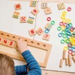 A child arranges numbered wooden tiles on a board, celebrating Pi Day with colorful rings, rods, and additional numbered tiles scattered on the table.