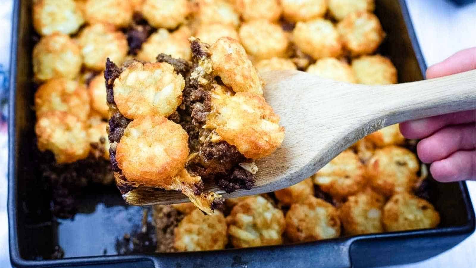 A wooden spatula lifts a serving of tater tot casserole from a baking dish filled with browned tots and ground beef.