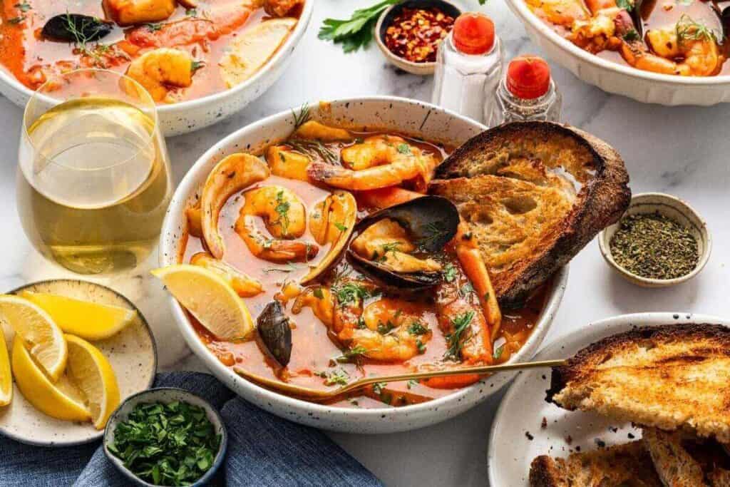 Bowl of seafood stew with shrimp, mussels, and herbs, served with toasted bread, lemon wedges, chopped parsley, a glass of white wine, and seasonings on a table.