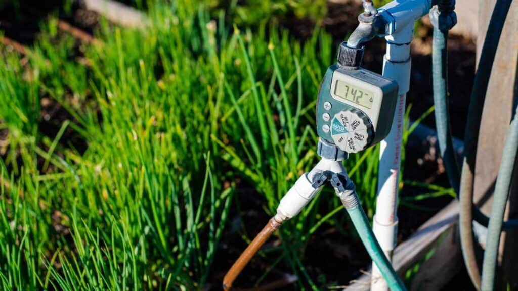 A digital water timer is attached to garden irrigation pipes, with green grass growing nearby.