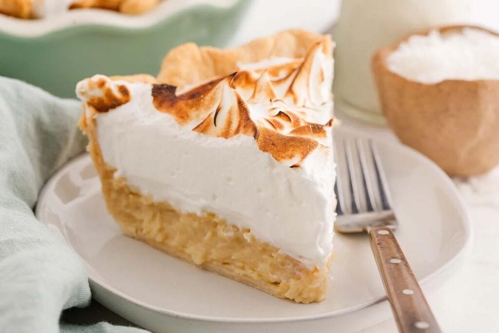 A slice of coconut cream pie with toasted meringue topping sits on a white plate next to a fork.